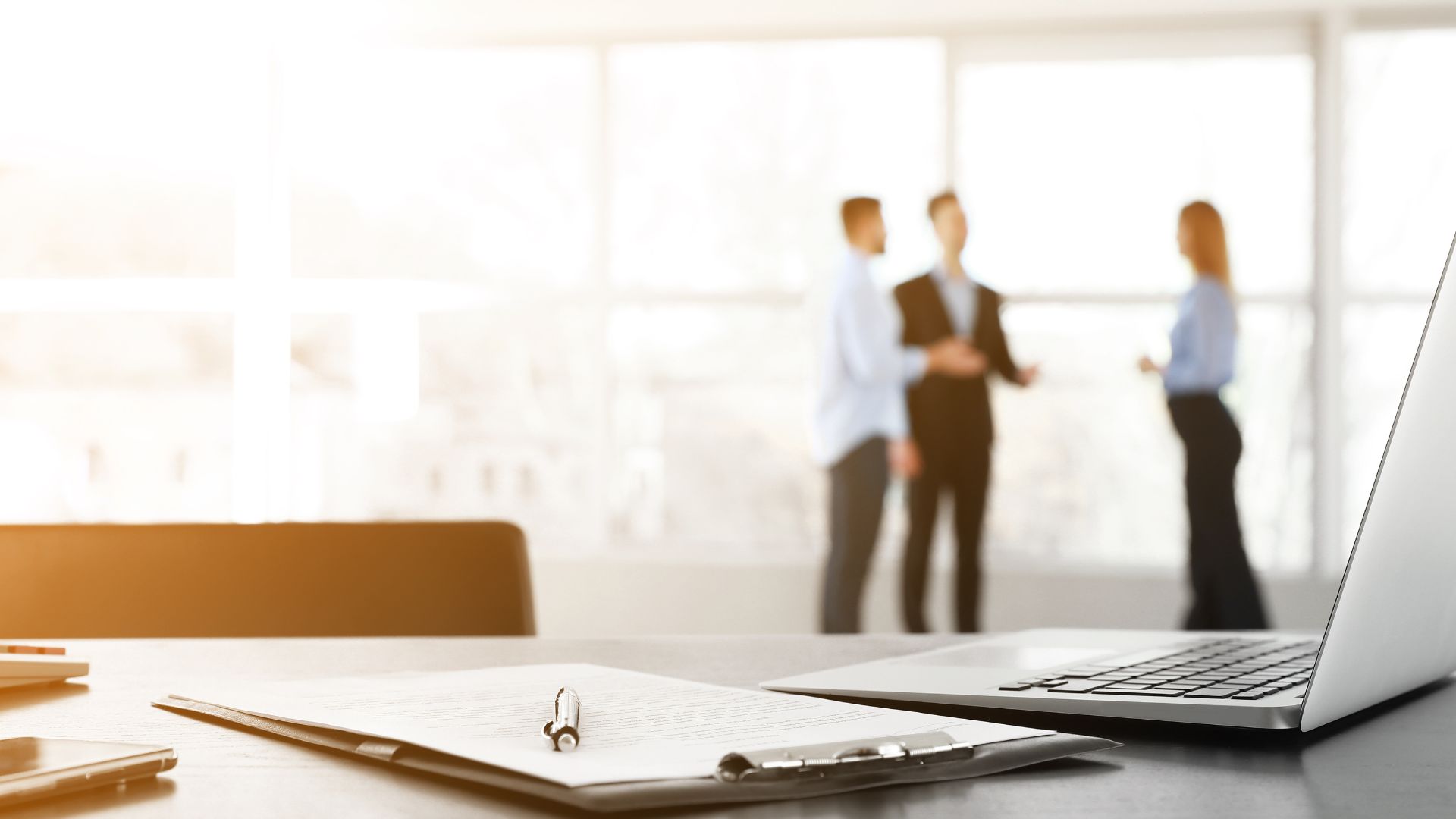 Business meeting scene with laptop, clipboard, and blurred colleagues